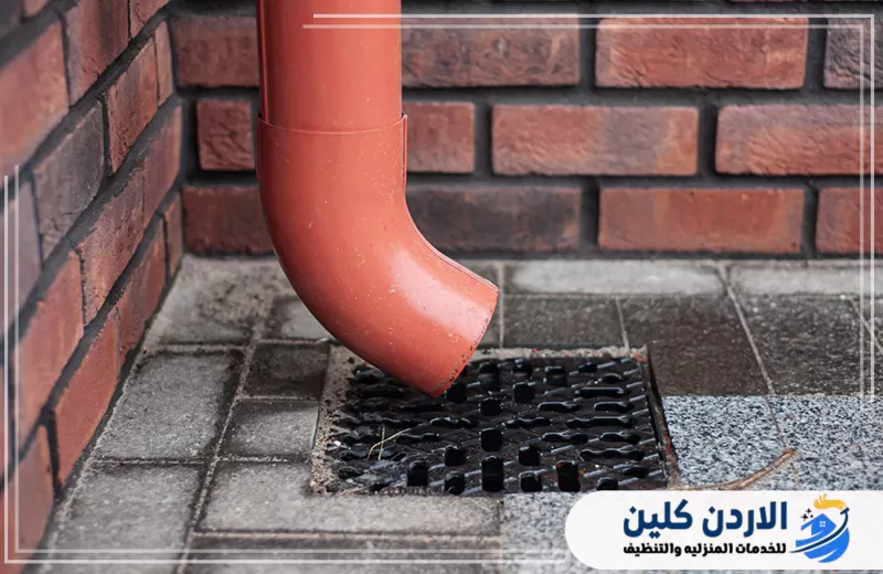 اطلب تركيب صرف أمطار ومزاريب عبر 0798779667. ماسورة صرف أمطار باللون الأحمر متصلة بفتحة تصريف أرضية لتوجيه المياه بعيدًا عن الجدار.