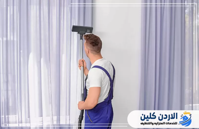 احجز تنظيف الستائر الطويلة مع الأردن كلين 0798779667. عامل يستخدم جهاز شفط لتنظيف الستائر الطويلة وإزالة الغبار من القماش بشكل منظم.