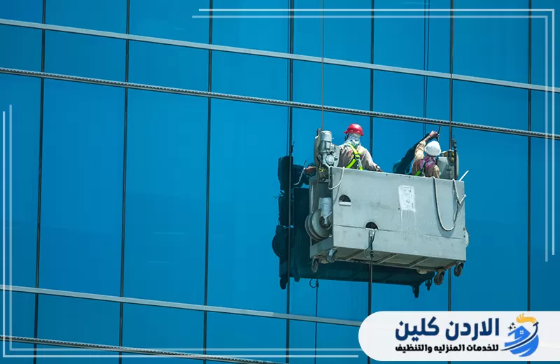 اطلب تنظيف الواجهات الزجاجية من الأردن كلين 0798779667. فريق متخصص ينظف واجهة زجاجية زرقاء من منصة معلقة مع استخدام أدوات وتجهيزات مناسبة.