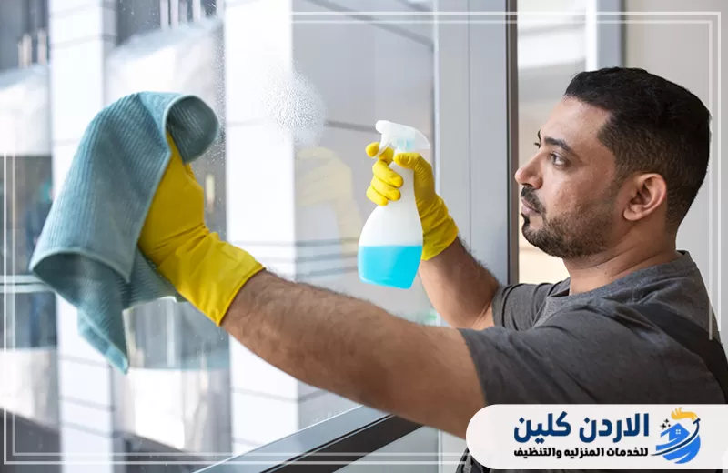 عامل ينظف زجاج النافذة باستخدام بخاخ وقطعة مايكروفايبر لإزالة البقع وآثار الأتربة.