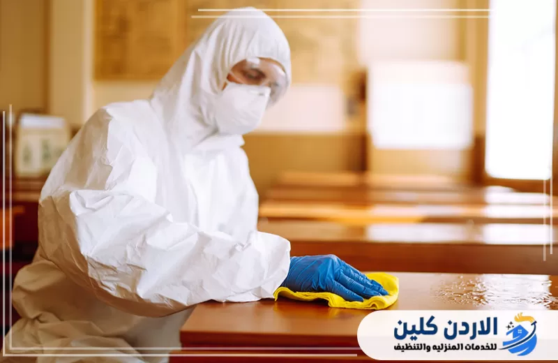 عامل يرتدي ملابس وقاية ويعقم الطاولات داخل الفصل للمساعدة في الحفاظ على نظافة البيئة التعليمية.