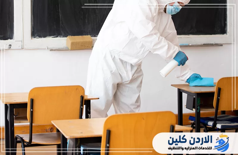 فني يعقم طاولات الطلاب داخل المدرسة باستخدام بخاخ وقطعة تنظيف مناسبة للأسطح.