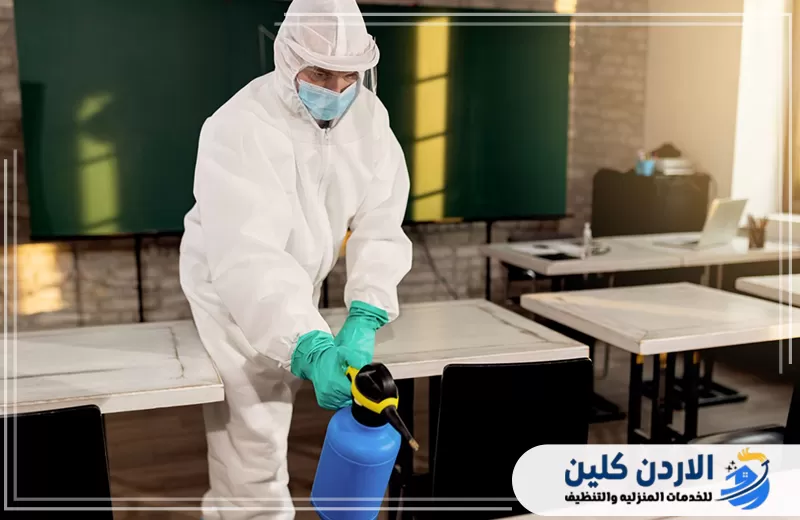 عامل يرتدي ملابس وقاية ويقوم بتعقيم الطاولات داخل الفصل للحد من انتقال الأوساخ والملوثات.