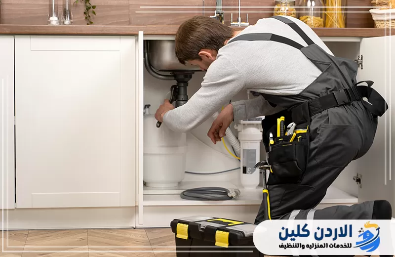 فني يفحص التمديدات أسفل حوض المطبخ ويجري صيانة لوصلات الصرف والمياه داخل الخزانة السفلية.